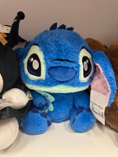 This is a close-up, top-down shot of a blue plush toy, likely a character from a Disney movie, possibly Stitch. The toy is positioned diagonally, with its head taking up most of the frame. It has large, oval eyes with black pupils and white highlights. Its ears are visible, with pink inner lining. A white tag is attached to one of the ears, with Polish text on it. The toy appears soft and furry. To the right of the blue toy, a paw of the same toy is visible. In the background, a brown furry object, possibly another stuffed animal, is partially visible. At the bottom of the frame, parts of other stuffed toys are visible, including a black and white one, and a white fluffy one wrapped in a sheer, sparkly netting. The lighting suggests an indoor setting, possibly a store display or a collection of toys. The image does not convey any specific time of day, weather conditions, or strong emotions. The visible text on the tag reads "Pluszowe zabawki" which translates to "Plush toys" in Polish, and a price of "8.00" is also visible. There are no people visible in the image. The context of Keila, Estonia is provided but not visually evident in the image itself.