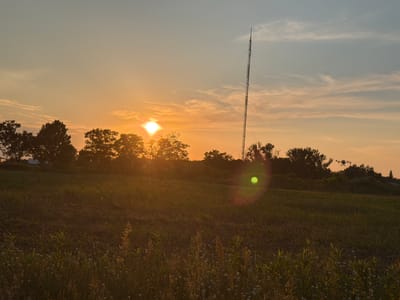 The image captures a sunset over a rural landscape, likely in or near the city of Łazy, Poland. The sun, a bright orb, is low on the horizon, casting a warm, orange glow across the sky and illuminating a tall, lattice-like radio tower positioned to the right of the frame. In the foreground, a field of tall grass and wildflowers is visible, with the light catching the tops of the plants. Silhouetted trees line the horizon, and in the distance, a small building or structure can be faintly seen. A drone is visible flying in the sky near the radio tower. The overall atmosphere is serene and peaceful, characteristic of a rural sunset.