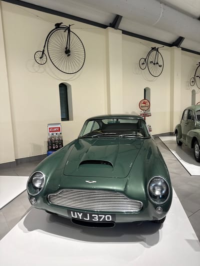 The image shows an indoor display featuring a green Aston Martin car. The car is parked on a white surface and is the main focal point. On the wall behind the car, there are several decorative vintage bicycles hanging. A vintage "Mobil Oil" sign and an "Atlantic" gas pump are also part of the display. The specific location cannot be determined from the image. No people are visible in the photograph. The environment appears to be a museum or showroom.