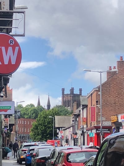 The image shows a street scene in Chester, United Kingdom, on a cloudy day. In the foreground, several cars are parked or moving slowly in traffic. A red circular sign with a white "W" and a registered trademark symbol is visible on the left side of the frame. Further down the street, there are shops with various signs, including "BG BAKERY" and "Traditional Home Cooking." In the background, the spires of Chester Cathedral are visible above the buildings and trees. The architecture suggests a historic city center. The overall impression is of a busy urban environment with a mix of modern vehicles and older buildings, characteristic of Chester.