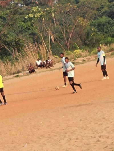 A group of people dressed in light-colored shirts and dark shorts are seen on a dirt field next to a wooded area. Some are actively playing what appears to be soccer. Other people are watching from the sidelines. One person in the group appears to be kicking a soccer ball.