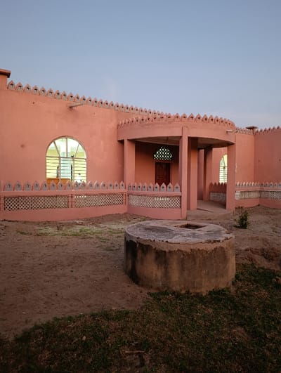The image depicts a salmon-pink colored building with a distinct architectural style, located in a courtyard of dry earth and sparse grass under a clear, light blue sky, in Vanga ward, Kenya. The building's roofline features a decorative parapet with repeating, pointed battlements, each adorned with a dark, cross-like emblem. A prominent curved portico, supported by two cylindrical pillars, frames a dark wooden door with an ornate, light-colored overhead mosaic. An arched window on the left side of the building exhibits interior illumination, revealing a light-colored frame and dark vertical bars. A low, decorative fence or railing, matching the building's color, runs along its base, featuring repeating white geometric cutouts. In the foreground, a weathered, circular concrete structure, identified as a well or cistern, is centrally positioned with a rectangular opening at its top. Patches of sparse green and brown grass are scattered across the unpaved ground, and a small bush is visible near the building's foundation to the right. No individuals are present within the frame.