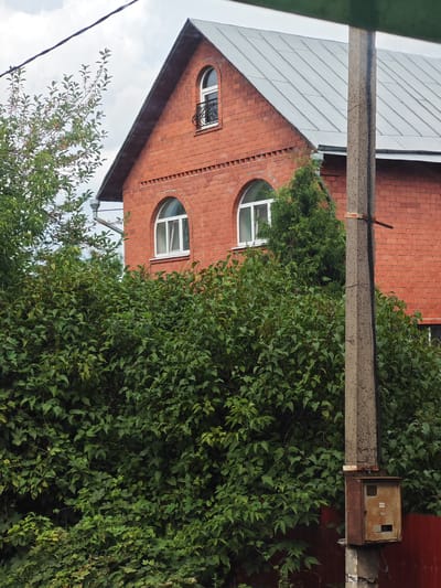 The image captures a close-up view of a red brick building with a gray metal roof and arched windows, partially obscured by dense green foliage. To the right, a concrete utility pole with a rusty electrical box is prominently featured. The scene appears to be in an outdoor setting, possibly a residential area in the village of Кабаново, Russia, given the presence of a typical utility pole and building materials. The sky is overcast. There are no people or animals visible in the frame.