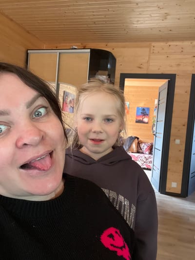 The image depicts a selfie taken indoors, featuring a woman and a young girl. The woman is in the foreground, sticking out her tongue with a playful expression. The girl is positioned behind her, smiling at the camera. The background shows a room with wood-paneled walls and a doorway leading to another room. Artwork is visible on the walls in the adjacent room. The location appears to be in a residence, possibly in Bol'shakino, Russia, based on external information.