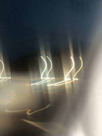 The image displays an abstract, motion-blurred night scene in South Oxfordshire, United Kingdom. The foreground and midground are dominated by several prominent, elongated streaks of bright white and yellow light against a dark blue-black background. Multiple vertical and upward-curving light trails exhibit distinct wavy or segmented patterns, suggesting artificial light sources in motion or a long exposure. A less intense, horizontal, wavy golden-yellow streak is visible in the lower-central area. The light trails emit a subtle glow and display minor chromatic aberration. No individuals, discernible stationary objects, or specific actions are visible; the scene consists primarily of illuminated light streaks within a dark environment.