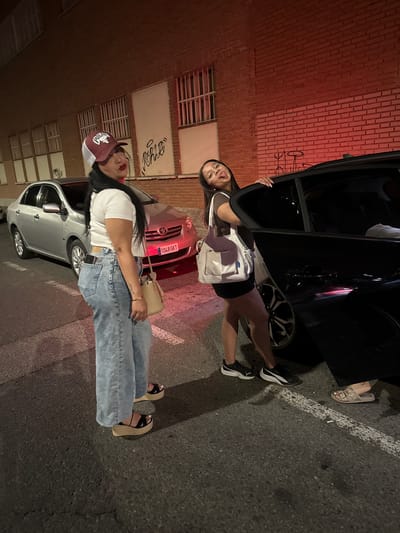 Two young women are standing on a street in Torrejón de Ardoz, Spain, next to parked cars at night. The woman on the left is wearing a white t-shirt, light blue wide-leg jeans, and platform sandals. She has dark hair, a maroon and white baseball cap, and is holding a beige purse. The woman on the right is wearing a dark top, a black skirt, and black sneakers. She has dark hair, glasses, and is sticking her tongue out as she leans on the open door of a black car. A silver car with a Spanish license plate reading "1048 GKT" is parked behind the woman on the left. The background features a brick building with windows and graffiti. The scene is illuminated by artificial lighting, casting shadows on the asphalt.