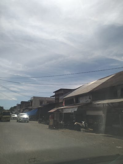 This image captures a street scene in Kruengmanyang, Indonesia, on a cloudy day. The sky is filled with wispy, light-colored clouds against a pale blue background, suggesting either early morning or late afternoon light. The foreground shows a paved road with a slight texture, indicating it might be a bit worn.

Along the right side of the road, a row of buildings with corrugated metal roofs lines the street. These buildings appear to be small shops or businesses, some with open fronts and awnings. One building has a sign with text that is partially obscured but includes what looks like "PHOTOCOPY & SPA" in English, and further text below in what appears to be Indonesian. Another sign reads "Sufiah". There are several parked motorcycles and what looks like some stacked materials outside one of the shops.

On the left side of the road, several vehicles are visible. A yellow truck is in the distance, followed by two white cars. The car closest to the viewer is a modern SUV, with its headlights on. The presence of these vehicles and the road suggest active local transportation.

The overall atmosphere is that of a typical day in a small town or village, with commerce and daily life unfolding. The weather seems mild, with the clouds diffusing the sunlight. There are no people prominently featured in the image, but the activity of vehicles and businesses implies human presence and ongoing routines. The setting is distinctly Indonesian, with the architectural style of the buildings and the presence of a truck commonly seen in Southeast Asia.