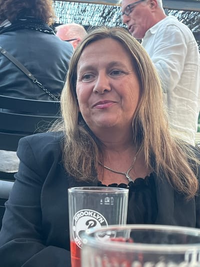 The image shows a medium shot of a woman in the foreground, with two other individuals partially visible in the background. The woman is looking to her left with a slight smile. She has medium-length, light brown hair and is wearing a dark blazer over a black top with a ruffled neckline and a diamond necklace. In front of her, on a table, are two glasses; one is a clear glass with the "BROOKLYN" logo and the other is a partially obscured glass filled with a red liquid. The background suggests an outdoor setting, possibly a restaurant or bar, with a thatched or woven ceiling overhead. A man wearing glasses and a white shirt is visible behind the woman. Another person with curly brown hair and a black garment with a white polka dot scarf is seated to the left, facing away from the camera. The context of Saint-Priest-en-Jarez, France is not visually supported by the image.