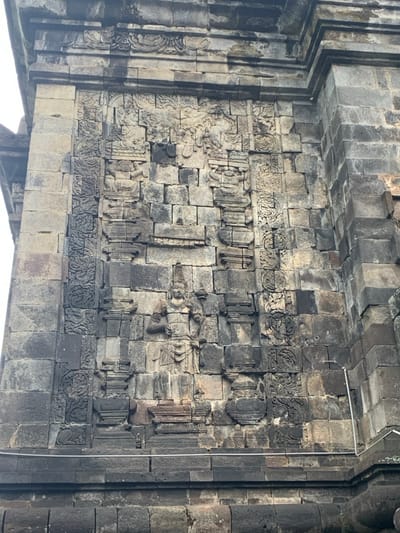 The image shows a close-up view of a stone wall with intricate carvings. The wall is made of gray stone blocks, some of which are missing or damaged. The carvings depict various figures and designs, including a prominent figure of a deity with a crown, standing in a pose of prayer. The wall appears to be part of a temple complex. The scene is located at latitude -7.605 and longitude 110.23, in the city of Mungkid, Indonesia. There are no people visible in the image. The carvings are likely from the ancient Hindu-Buddhist era, suggesting the location is a site of historical and cultural significance. The wall is weathered, displaying the effects of time and the elements. A metal wire runs across the bottom of the image.