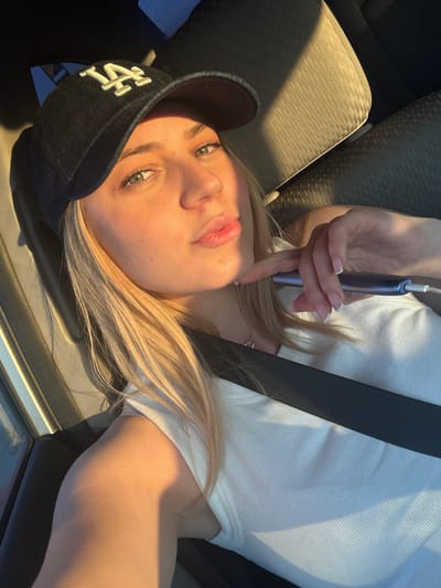 A person, seemingly female, is reclining in what appears to be a car seat. The person is wearing a black baseball cap with the letters "LA" on the front, a white t-shirt, and a seatbelt. The person is holding a blue electronic device near their face. The interior of the vehicle appears to be gray and black. The location is unknown and cannot be determined from this image. There is no information that can confirm the location to be Oakland, United States of America.