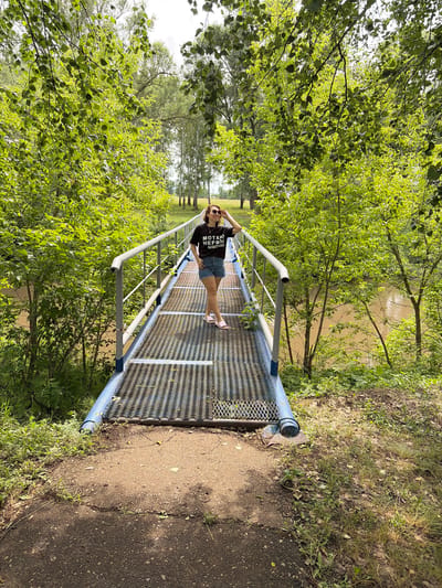 A full-shot image shows a woman standing on a pedestrian bridge over a body of water. The woman is wearing a black t-shirt with white lettering, denim shorts, and pink sandals. She has sunglasses on and her right hand is touching her hair. The bridge is made of metal with a patterned surface and has blue railings. The bridge connects to a dirt path that leads into a wooded area. The trees are lush and green, with sunlight filtering through the leaves. The water is a murky brown color. In the background, more trees and a field can be seen under a cloudy sky. The overall scene depicts a natural outdoor setting, possibly a park or recreational area.