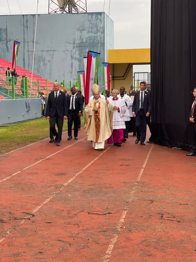 A wide, medium shot depicts a procession on a red running track, with several individuals, including a bishop and men in suits, moving towards the viewer.

The primary figure, a bishop in ornate gold and white vestments, holds a staff and appears to be the focus of the procession. He is flanked by individuals in dark suits and religious figures in white robes.

To the left, flags representing Equatorial Guinea are visible, along with a grey wall and stadium seating. To the right, the red running track extends into the distance. The ground is marked with faded white lines.

The background features a structure with yellow and grey sections, suggesting a building or stage area. Black curtains are partially visible in the top left corner.

The setting is likely a stadium or large outdoor venue in Malabo, Equatorial Guinea, given the presence of the national flags and the context of a formal procession.