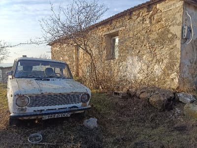A dilapidated white car sits in front of a weathered stone building. The car shows significant rust and decay, with a partially obscured license plate. The building, constructed of rough-hewn stones, has boarded-up windows and a decaying exterior. Sparse vegetation and bare branches surround the scene, suggesting a neglected or abandoned environment. A small electrical box is attached to the building's corner.