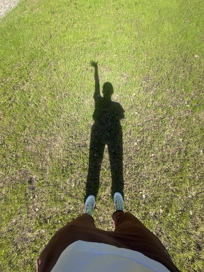 A ground-level view of a grassy field. The shadow of a person is visible on the grass, with their arm extended. Parts of the person are visible in the corner of the frame, including shoes and legs covered in brown pants. Location not explicitly stated.