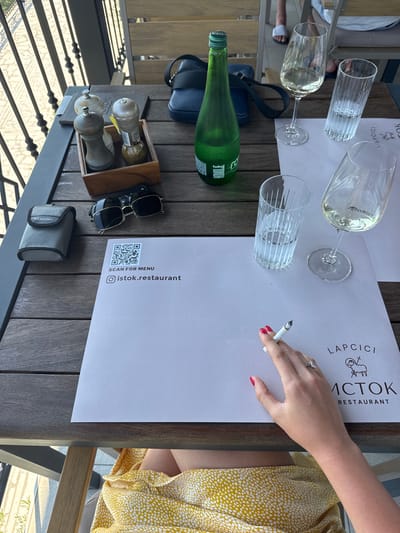 An outdoor table setting in Stanišići, Montenegro, features a wooden table with various objects. A woman's hand, with red-painted nails and a diamond ring, holds a lit cigarette over a paper placemat. The placemat has a QR code for a menu and the text "SCAN FOR MENU" and "@istok.restaurant". Next to it, a wine glass with white wine, an empty glass with water, and another empty wine glass are placed. In the background, a green glass bottle of soda, a blue bag, salt and pepper shakers in a wooden holder, a Samsung case, and sunglasses are visible. The table is situated on a balcony or terrace with a view of a railing and a sunlit cobblestone pathway.