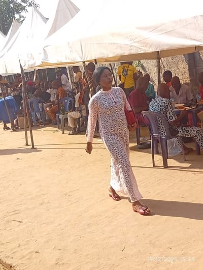 A woman in a long, white, patterned dress walks across a dirt-covered area. She wears red sandals. Behind her, under white canopies, a crowd of people sit in chairs around tables. Some are eating, and some are talking. A yellow shirt with an image on it is visible. The photo is taken in bright daylight. A date stamp "19/12/2025 15:34" is in the lower right corner.