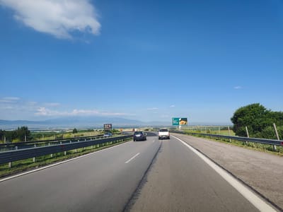 The image captures a highway scene on a clear day, with a blue sky and scattered clouds. The viewpoint is from inside a vehicle, looking forward along a multi-lane asphalt road. Two cars are visible in front, a dark blue hatchback and a white SUV. A metal guardrail runs along the right side of the road, separating it from a grassy verge and a line of trees and bushes beyond. In the distance, rolling hills and mountains form the horizon, with a hazy landscape of fields and structures in the valley. Several billboards are present along the roadside, one featuring a burger advertisement and another with a portrait and text. The scene is consistent with a highway outside a populated area, likely near the city of Eleshnitsa, Bulgaria, given the visual cues of the landscape and potentially the text on the billboards, although the text is not fully legible.