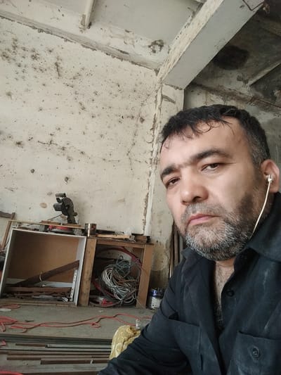 The image shows a man with short, dark hair and a beard wearing a black shirt. He is looking at the camera, and has a serious expression on his face. He is wearing a white earbud in his right ear. The background appears to be an indoor space, possibly a workshop or construction site, with a concrete wall, exposed pipes, and various tools and materials scattered around. There is a metal cutting saw visible on a shelf. The overall impression is of an industrial or unfinished environment. There is no specific indication of the city of Chalysh, Uzbekistan, in the image itself.