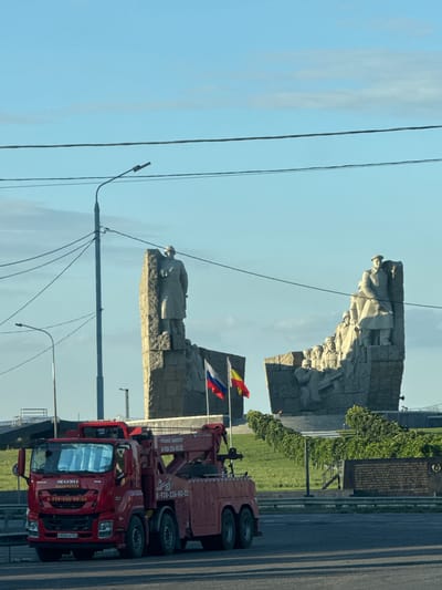 The image depicts the Sambek memorial complex in Sambek, Russia, under a clear blue sky with scattered clouds. In the foreground, a red ISUZU tow truck is parked on a road. The truck has visible branding, including "ISUZU" and "ABARYATOP," along with phone numbers and a license plate that reads "P856PB 761". Behind the truck, the monumental Sambek memorial complex is prominent. This complex features a large stone sculpture of soldiers. Two flags, one appearing to be the Russian flag and another with yellow and red bands, are flying near the memorial. The landscape surrounding the memorial is green and appears to be a grassy hillside. A dark stone plaque with inscriptions is situated at the base of the hill. Power lines and streetlights are visible in the background, along with a glimpse of a bridge or elevated structure. The overall scene is a daytime shot with clear visibility and no discernible human activity directly within the memorial complex itself.