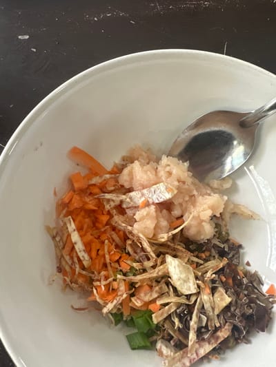 A white bowl holds a mix of food, including shredded carrots, chopped green onions, and a light-colored, minced protein. Crispy fried taro strips are scattered throughout, adding texture. A portion of darker, finely chopped material, potentially dried mushrooms or seaweed, sits on top. A silver spoon is resting in the bowl, suggesting the food is ready to be eaten. The background is a dark, potentially speckled, surface.