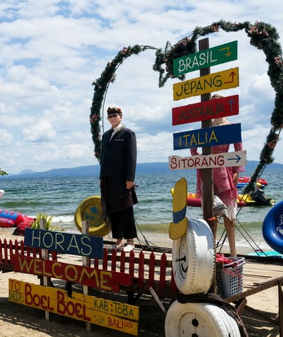 An outdoor, eye-level, full shot captures a man in traditional Indonesian attire standing by a heart-shaped archway adorned with flowers and greenery. He is positioned to the left of a directional signpost listing various global destinations, including Brasil, Jepang, Australia, Italia, and Sitorang. The man wears a dark suit, a patterned sarong, a white shirt, and a headscarf. Behind him, a woman in a pink dress stands partially obscured by the signpost. The scene is set on a beach with the ocean in the background, the water appearing calm with distant hills visible on the horizon. The beach is decorated with colorful inflatable toys and a red picket fence. A series of signs in the foreground indicate "HORAS WELCOME TO DESA: Boel Boel KAB: TOBA KUTA BALI GEEE". The sky is partly cloudy. The image appears to be taken in the city of Lumban Bul Bul, Indonesia, likely a tourist attraction or photo opportunity.