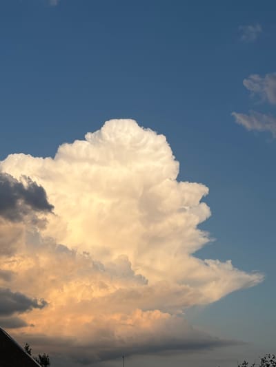 The image captures a dramatic sky scene over the city of Slobozia, Moldova. A large, towering cumulus cloud dominates the center of the frame, illuminated by warm, golden light from the setting sun, suggesting late afternoon or early evening. The cloud exhibits a bright white, fluffy upper portion, while its lower sections are tinged with shades of orange and yellow, with darker grey clouds present to the left. The background is a clear, deep blue sky, with a few smaller, wispy clouds scattered in the upper right portion. In the lower portion of the image, a dark, angled roofline of a building is visible on the left, and the tops of dark green trees can be seen along the bottom edge, indicating an urban or suburban setting within Slobozia. No people or specific actions are depicted. The overall impression is one of natural atmospheric beauty.