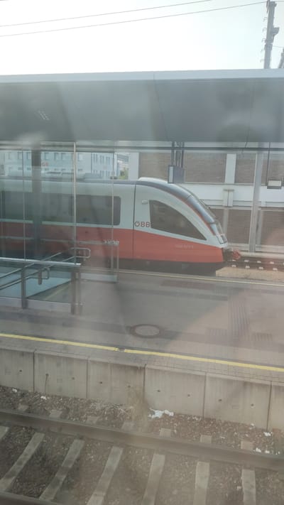 The image shows a modern red and white train at a station in Sankt Pölten, Austria. The train has the OBB logo and the number 5022 023-3 visible on its side. The station platform is visible, with a yellow line marking the edge. In the background, there are buildings and infrastructure typical of a train station, including a covered area over the platform. The image appears to be taken from inside another train, as indicated by the reflection on the glass. The scene is a daytime shot with a bright sky.