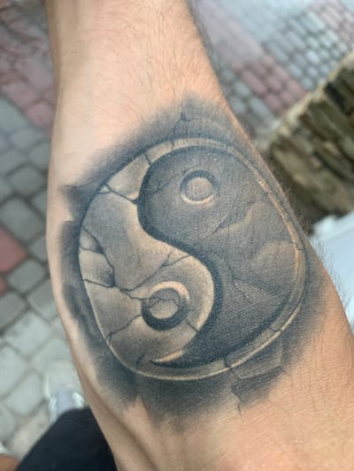 A tattoo of a cracked yin and yang symbol is visible on a person's arm. The tattoo is done in grayscale with shading to create a textured, stone-like appearance. The cracks on the symbol are rendered with fine black lines. The arm is positioned over a blurred background that appears to be an outdoor setting with cobblestones or a similar paved surface and some greenery or decorative elements. The image does not provide information about the city of Khust, Ukraine.