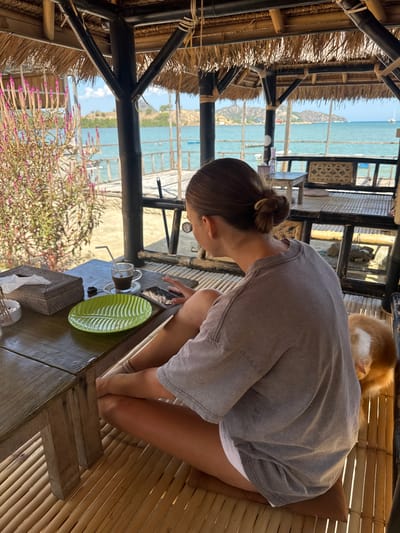 A woman with her hair pulled back in a bun sits cross-legged on a bamboo floor in an open-air pavilion. She is wearing a grey t-shirt and white shorts. In front of her on a wooden table is a cup of coffee with a straw, a green leaf-shaped plate, a tissue box, and a smartphone. Her left hand is resting on the phone. To her right, a ginger and white cat is visible. Through the open sides of the pavilion, a view of the ocean, a wooden pier, and a distant shoreline with hills can be seen. The pavilion has a thatched roof and bamboo supports. The scene appears to be located in Kertasari, Indonesia, likely at a beachfront establishment.