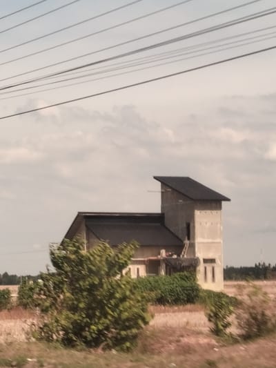 A two-story building stands in Keudebagok Dua, Indonesia. The building features a dark roof and unfinished exterior walls. It appears partially obscured by vegetation. Power lines stretch across the upper portion of the image. The surrounding landscape is open and dry.