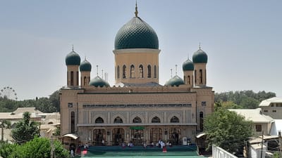 The image shows the Juma Mosque in Denov, Uzbekistan, a large, multi-domed mosque with tan stone walls and green tiled domes. The mosque is flanked by two tall minarets, also topped with green domes. The main dome is significantly larger and sits atop the central section of the building, which has a pale yellow facade with rows of arched windows. Intricate geometric patterns and Arabic calligraphy adorn the facade below the domes. In front of the mosque, a wide set of green stairs leads up to the entrance, where several people are seen sitting on prayer rugs. Some individuals are standing near the entrance, and one person is walking up the stairs. The surrounding area shows other buildings, lush green trees, and in the distance, a Ferris wheel is visible, suggesting an urban environment with recreational facilities. The sky is clear and pale blue, indicating a sunny day.