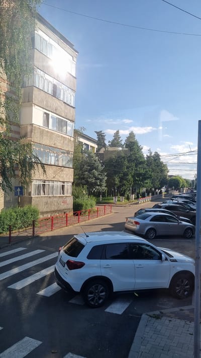 A white Suzuki Vitara is parked on a pedestrian crossing. The vehicle is oriented diagonally, with its rear on the right side of the frame and its front on the left. The pedestrian crossing consists of white stripes on dark asphalt. Adjacent to the crossing is a sidewalk with brick paving. In the background, a row of parked cars, including a silver Audi A4, are visible along the street. A building with concrete balconies and numerous windows is situated on the left side of the image, with trees and greenery in front of it. The scene appears to be in a residential area. The text "SECOURS" is faintly visible reflected on a window in the background, suggesting a potential presence or association with emergency services, possibly within the context of the city of Fălticeni, Romania, given the prompt's specification. The sky is blue with some scattered clouds, and the sunlight creates glare on the building's windows. A red metal fence runs along the edge of the sidewalk in front of the building.
