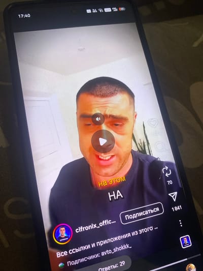 This media file shows a close-up of a smartphone screen displaying a social media video. The primary subject is a man's face, captured in a candid or humorous moment, with his mouth open in a smile or laugh. His expression suggests amusement or engagement with the video content.
The video interface is visible, indicating it's a short-form video platform, possibly TikTok or a similar application. Icons for likes (1628) and comments (2289) are present, along with a share icon (70) and a "Follow" button (Подписаться) next to the number 1841. A profile picture of the man, labeled "CIFRONIX," is also visible.
Text overlays on the video include "на этом" (on this) and "HA" in Russian, possibly part of a caption or a spoken phrase. Below the video, further Russian text reads "Все ссылки и приложения из этого ..." (All links and applications from this...) and "Подписчики: avto_shokkk_" (Subscribers: avto_shokkk_), and "Ответы: 29" (Answers: 29).
The phone itself is resting on a textured surface, likely a fabric like a blanket or sofa, in a dimly lit indoor setting. The time displayed on the phone's status bar is 17:40, suggesting it's late afternoon. The phone's signal indicators (VPN, 4G, VOLTE) are also visible. The overall context suggests the person is using their phone to watch or record a video.