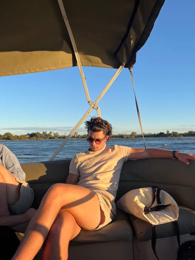 A man with sunglasses is reclining on a boat, likely on the Zambezi River near Victoria Falls, Zimbabwe. The river and its shoreline are visible in the background. The boat has a shade canopy and beige seating with a white bag beside the man. A second person is partly visible near the bottom of the frame.