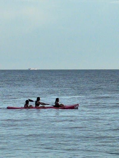 Three individuals are in a red kayak on a body of water. The kayak is positioned horizontally in the frame, with the individuals facing towards the left. The person in the front left appears to be a child, with their back to the camera. The person in the middle is an adult, also facing left, holding a paddle and appearing to row. The third person, in the rear right, is partially visible and also facing left. The water is blue and appears slightly choppy. In the distance, a large ship or barge is visible on the horizon, situated between the water and a pale blue sky. The scene is set outdoors, likely in the ocean, with no visible urban structures or city landmarks of Dauin, Philippines.