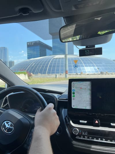 The image, taken from inside a Toyota vehicle, captures a view of the Spodek arena in Katowice, Poland, under a clear blue sky. A person's hand is visible on the steering wheel, indicating they are driving. The car's dashboard displays include a navigation system showing a map of Katowice and a digital speedometer reading 52 km/h, alongside a Navitel dashcam. In the background, modern glass buildings stand out against the sky, with the distinctive geodesic dome of Spodek prominently featured. The scene appears to be set on a sunny day, with green grass and foliage visible in the foreground.
