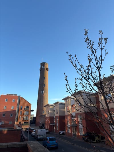 The image depicts a view in Chester, United Kingdom, capturing a tall, brick-built cylindrical tower, likely a former industrial chimney or similar structure. The tower is positioned in the mid-ground, under a clear blue sky with some bare branches and emerging buds of a tree obscuring part of the top right. The architecture around the tower appears to be residential and commercial buildings with a mix of red brick and light-colored facades. A street runs alongside the tower, featuring parked vehicles. The scene conveys a sunny, urban environment.