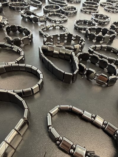 A close-up photograph shows an array of magnetic hematite bracelets displayed on a flat, dark surface. The bracelets vary in design, with some consisting of rectangular beads separated by spherical beads, and others using hexagonal or square beads. The bracelets are arranged in a seemingly random pattern, partially overlapping each other. The bracelets' surfaces reflect light, giving them a metallic sheen. The background is a plain, dark gray surface, possibly a display case or table.
I don't have information about the bracelets being in the city of Синьково, Russia.