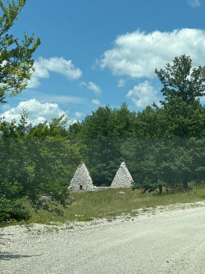 The image depicts two conical stone structures situated in a grassy field, surrounded by dense green trees under a bright blue sky with scattered white clouds. A gravel road runs along the bottom right of the frame. The structures appear to be ancient ruins, with one of them showing a small, dark rectangular opening. The environment suggests a natural, undeveloped landscape, likely in a rural or park setting. This scene is located near the city of Nikšić, Montenegro.