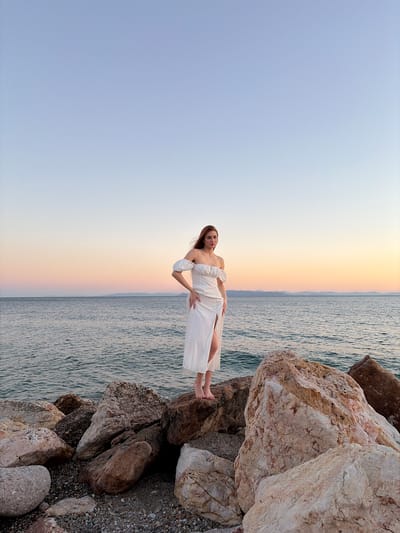 A young woman with reddish-brown hair stands on a rocky shoreline in Paleo Faliro, Greece. She is wearing a white, off-the-shoulder maxi dress with a high slit. The woman is facing the camera, with her left hand on her hip and her right arm bent at the elbow. The sea is calm and extends to the horizon, where a gentle gradient of colors indicates either sunrise or sunset. The sky is clear, transitioning from a pale blue at the top to softer hues of pink and orange near the horizon. The foreground is dominated by large, uneven rocks of various sizes and textures, some appearing smooth and rounded, others rough and angular. Small pebbles and gravel are visible between the larger rocks. The overall scene conveys a sense of tranquility and natural beauty.