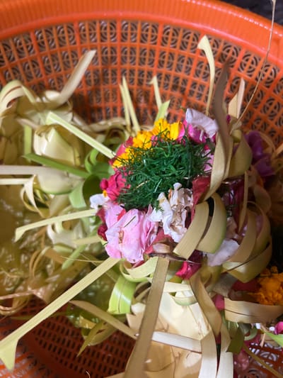 A close-up overhead shot shows a woven orange plastic basket containing a floral offering. The basket has a grid pattern with square openings. Inside the basket are various flowers in shades of pink, white, yellow, and purple, mixed with green foliage and thin, pale green palm fronds that are woven into decorative shapes. Some of the palm fronds are curled and twisted, wrapping around the flowers. The offering appears to be placed on a bed of more palm fronds, which extend out from the center. The lighting is bright, creating some highlights and shadows on the floral arrangement. The image was taken in Mendoyo, Indonesia, where such offerings are common.