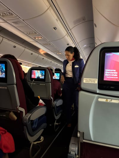 The image depicts the interior of an airplane cabin, likely in flight. Rows of maroon-colored seats are visible, equipped with individual entertainment screens. Passengers are seated, with at least one flight attendant standing in the aisle. The screens display a welcome message in multiple languages (English, Spanish, and Portuguese). Overhead compartments and individual passenger controls are visible along the cabin wall. Text on a nearby seat indicates the plane model is a "767-300". While the airline is not specified, the screen in the front says "LATAM PLAY". Without more context, the location in Pudahuel, Chile is difficult to confirm.