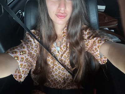 A young woman with long, dark brown hair is sitting in the driver's seat of a car, wearing a pink, orange, and yellow patterned button-down shirt. She is looking towards the camera with her lips pursed. A black seatbelt is visible across her chest. The car's interior is dark, with black leather seats and a glimpse of a brown purse on the passenger seat. The image was taken in Deerfield Beach, United States of America.