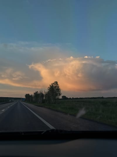 The image depicts a view from inside a vehicle, likely on a highway, in Voskresensky District, Russia. The road stretches into the distance with a single white line marking lanes. A field borders the road on one side, and trees are visible in the mid-ground. The sky is mostly blue with large, illuminated clouds that appear to be sunset or sunrise colors, probably from sunlight. A portion of the vehicle's interior is seen in the bottom-right corner.