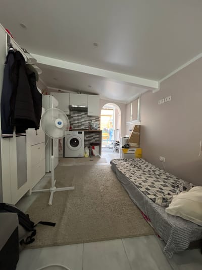 The image depicts the interior of a compact living space, likely a studio apartment, located in Stains, France. The room is furnished with essential items, suggesting a functional living arrangement.
On the left side of the room, a white wardrobe or storage unit is visible, with several dark-colored jackets hanging on a rack. Adjacent to this is a white standing fan with a blue and silver emblem on its center. Further into the room, on the left, is a small kitchen area. This area includes white cabinets, a tiled backsplash, a washing machine, and a countertop with a kettle and other kitchen items.
The central and right portions of the image are dominated by a floor-level bed with a patterned duvet and a white pillow. A beige rug covers a portion of the floor in front of the kitchen area and extends towards the bed. On the wall above the bed, there are three electrical outlets. To the right of the bed, there are stacked cardboard boxes and other items.
The overall environment is utilitarian, with a mix of storage solutions and living necessities. The space appears somewhat cluttered, but functional. The natural light enters from an arched doorway at the far end of the room, which leads to an outdoor area. The flooring is a combination of what appears to be light-colored tiles and the rug. The walls are painted a pale pinkish-beige.