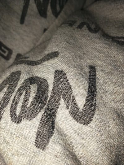 The image is a close-up, slightly blurry shot of a grey fabric with black lettering. The fabric appears to be a soft material, possibly cotton, with a visible knit texture. The black lettering is stylized and appears to form words, though they are not entirely legible due to the angle and focus. Some letters are clearly visible as "M" and "N", with other indistinct shapes suggesting more letters. The scene is likely indoors, given the lighting and focus on the fabric. The location in Rumuomasi, Nigeria, is a residential or commercial area, but the image itself does not provide any specific contextual clues about the city.