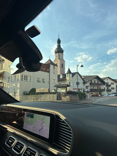 This media file captures a view from inside a car, looking out through the windshield and side window. The scene is a charming European town, likely in Italy given the location context of Kastelruth - Castelrotto. Dominating the background is a tall, ornate church tower with a pointed spire and a clock face, characteristic of historic European architecture. Adjacent to the church are several buildings with traditional pitched roofs, some featuring balconies adorned with flowers. A street with a pedestrian crossing is visible, and a few people are walking along the sidewalk.
A sign reading "Saalstuben" is prominent on one of the buildings, suggesting a restaurant or accommodation. Another sign with "EURO" is also visible. Inside the car, the dashboard is partially visible, including a digital screen displaying a navigation map with a route marked in blue. The time is indicated as 14.8 minutes to destination and the time as 20:53.
The sky is a bright blue with scattered white clouds, indicating fair weather. The lighting suggests it is late afternoon or early evening, with the sun casting long shadows. The overall impression is one of a peaceful and picturesque town, viewed from the perspective of someone traveling through it. The image is oriented vertically, implying it was taken with the phone held upright.