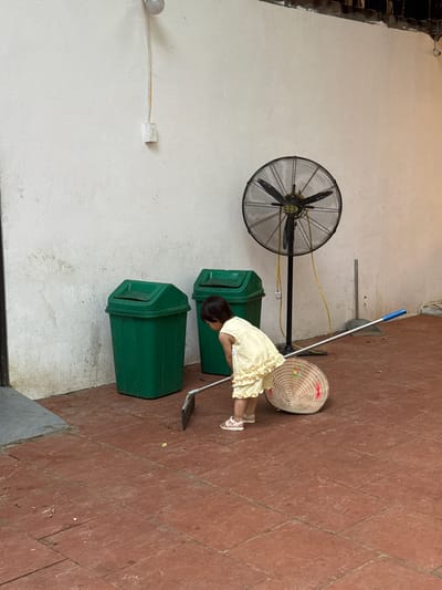A young child in a yellow dress and sandals is sweeping a tiled floor with a broom. A traditional conical hat lies on its side on the floor near the child. In the background, two green trash cans stand against a white wall. A large industrial fan on a stand is also visible, along with a dustpan and broom leaning against the wall. The scene appears to be an outdoor covered area, possibly a patio or walkway. The location is identified as Cổ Lũng Commune, Vietnam.