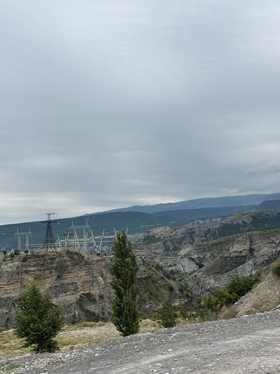 This is a photograph of the Chirkey Hydroelectric Power Station in Chirkey, Russia. The power station is situated in a mountainous, rugged landscape. In the foreground, a gravel road leads the viewer's eye towards a vast, layered rock formation on the left side of the image. Two trees, one smaller and bushier on the left and a taller, slender one on the right, stand in front of the rock face.
In the midground, the imposing structures of the Chirkey Hydroelectric Power Station are visible. Large power lines supported by tall metal towers stretch across the scene. The station appears to be built into the side of the mountain. The background reveals a series of rolling hills and mountains, covered in vegetation and shrouded in a hazy, overcast sky. The overall impression is one of industrial infrastructure integrated into a dramatic natural setting.