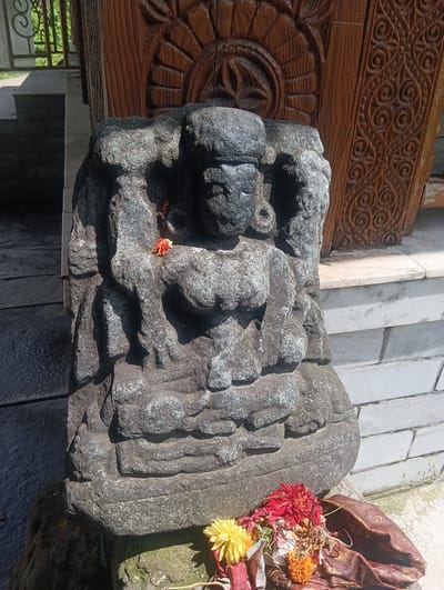 A stone sculpture of a multi-armed deity is the central focus of this image, set against a backdrop of ornate wooden carvings and a white brick wall. The sculpture, depicting a seated figure with visible torsos and limbs, is weathered and dark gray, with some floral offerings at its base. Red, yellow, and orange flowers are arranged in a pile, partially covered by a dark, shimmering fabric. The deity sculpture is placed on a stone platform, with a paved area in the foreground and a step leading to a shaded porch area in the background. The scene appears to be an outdoor religious or historical site. No people are visible in the image. The location is identified as Gojra, India, though specific details about the site within Gojra are not provided.
